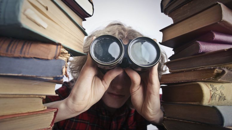 anonymous person with binoculars looking through stacked books